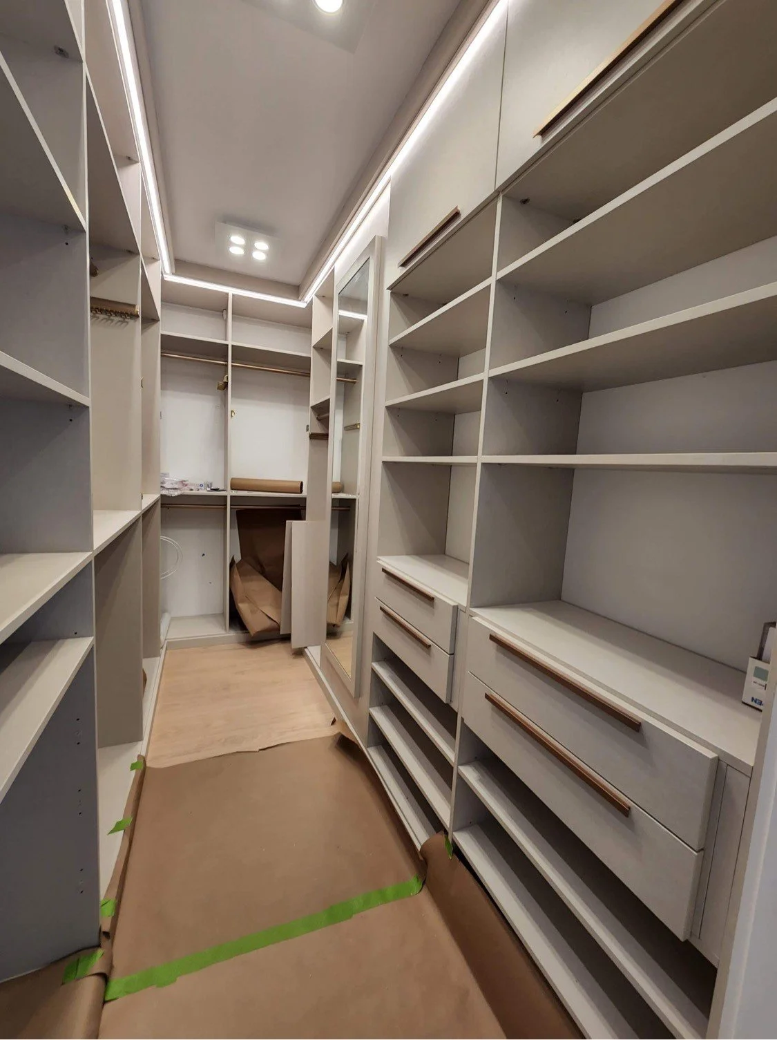 An empty walk-in closet with white shelves, drawers, and hanging rods. Protective brown paper covers the floor and some shelves, and the lighting is bright.