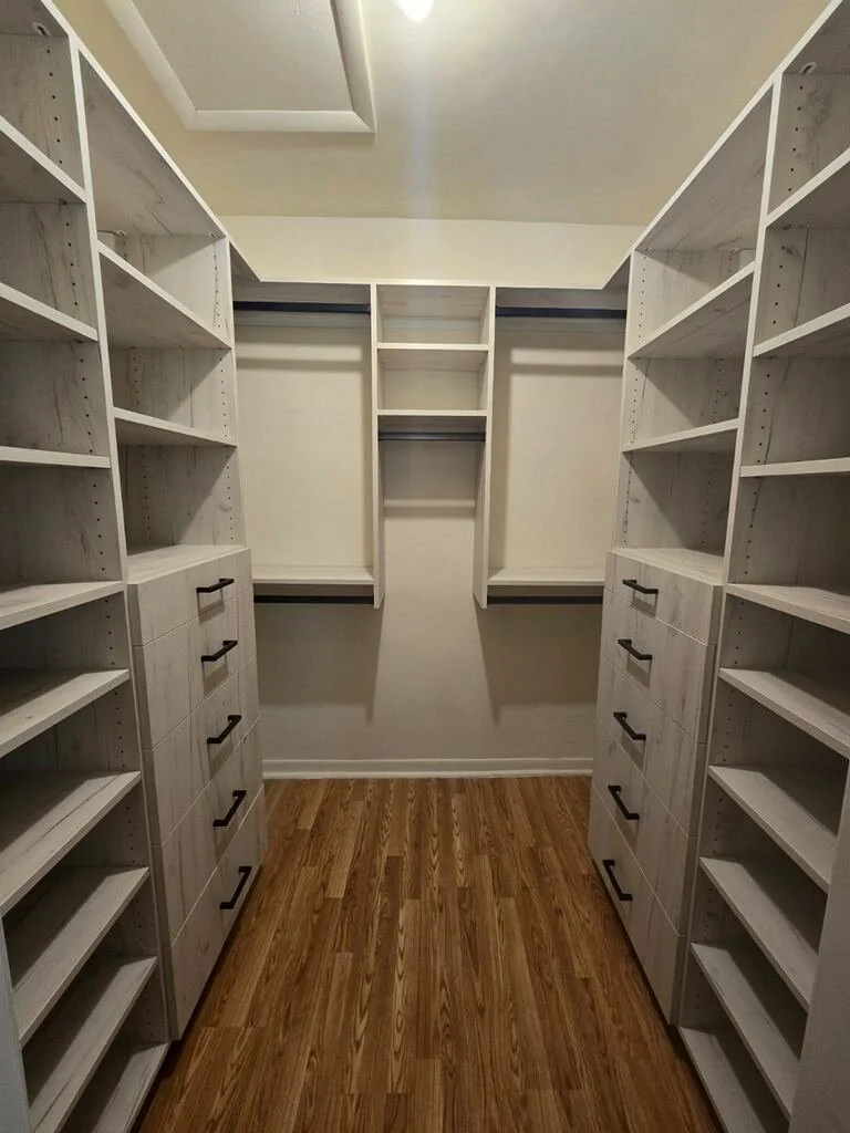 Empty walk-in closet with white built-in shelves, drawers, and hanging rods, hardwood floor, and a small skylight.