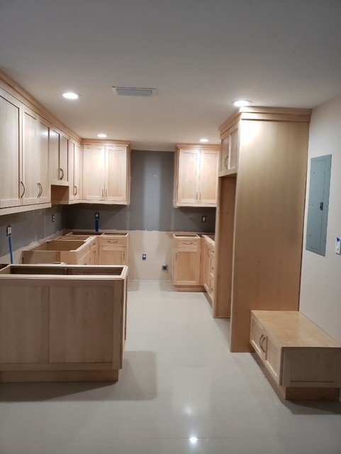 Empty kitchen with light wood cabinets, some upper and lower, and a small bench or storage outside the kitchen area.
