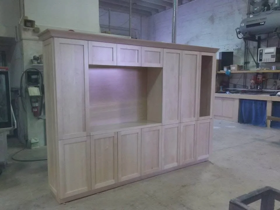Unfinished wooden cabinet in a workshop with shelves, tools, and equipment behind it.
