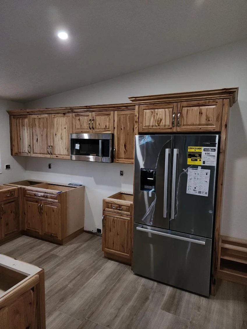 Kitchen with unfinished wooden cabinets, a stainless steel side-by-side refrigerator, a microwave, and empty spaces for appliances or sinks. The flooring is wooden and the walls are plain white.