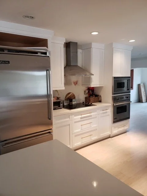 Modern kitchen with white cabinetry, stainless steel appliances, including a large refrigerator, microwave, and oven, with a stainless steel range hood, and light-colored hardwood floors.