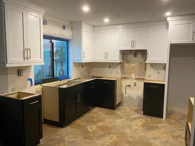 Kitchen under renovation with white upper cabinets, black lower cabinets, and a window showing outdoor plants.