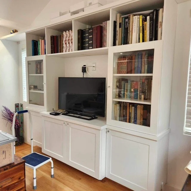 White built-in bookshelf unit with books and a TV in the center, a small blue and white chair in front, and a wooden floor.