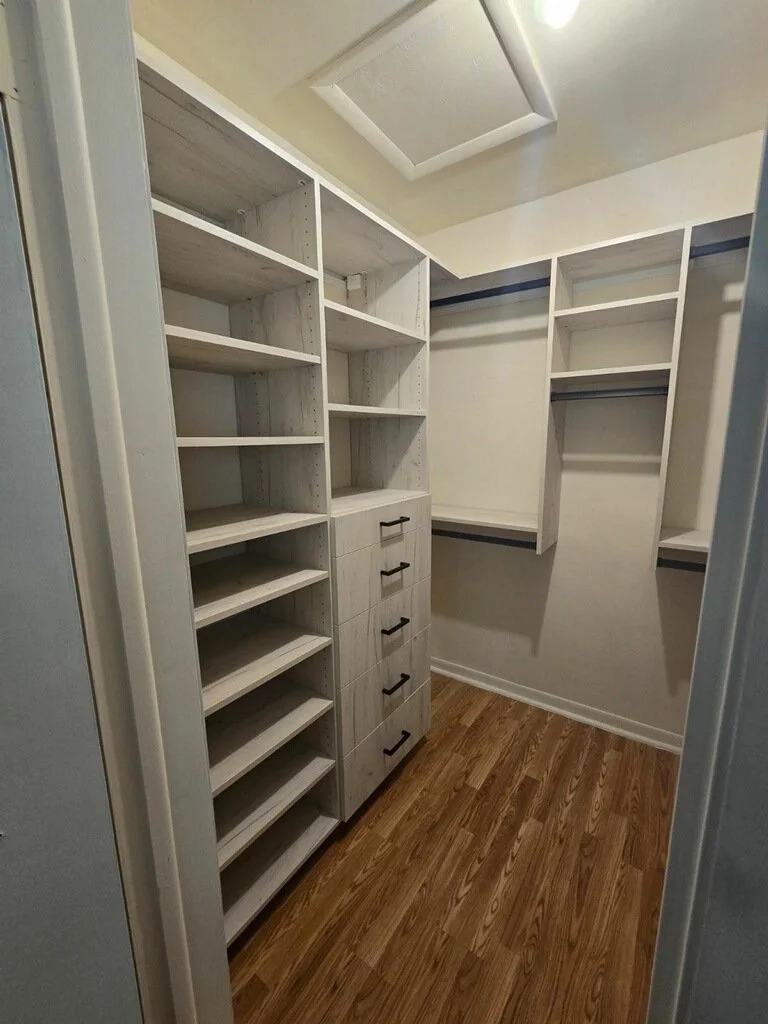 Empty walk-in closet with white wooden shelves, drawers, and hanging rods on a wooden floor.