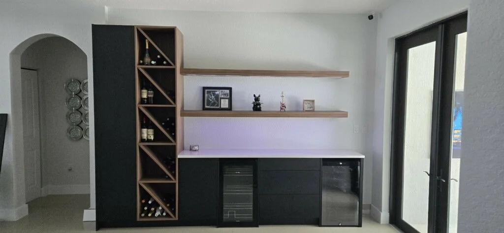 Modern kitchen bar area with built-in wine rack, open wooden shelves, and a white countertop. There are wine bottles in the rack and decorative items on the shelves. Opposite a glass door that leads outside.