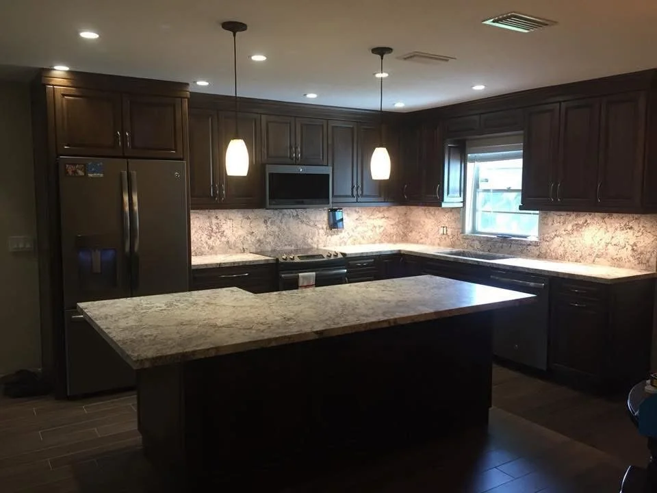 Modern kitchen with dark wooden cabinets, granite countertops, stainless steel refrigerator, microwave, and stove, illuminated by recessed lights and hanging pendant lights.