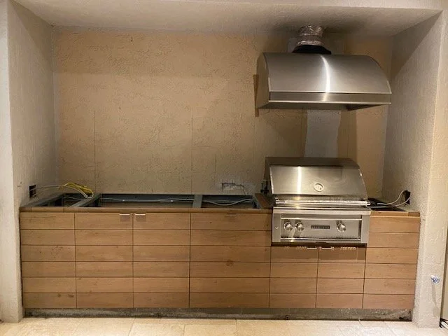 An outdoor kitchen area with a built-in grill, a range hood, and wooden paneling, but the grill and other appliances are missing.