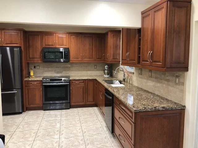 Kitchen with wooden cabinets, granite countertop, stainless steel appliances including a refrigerator, microwave, oven, and dishwasher, beige tile floor, and a small window above the sink.
