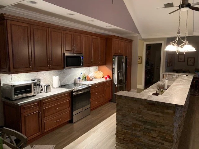 Kitchen with wooden cabinets, marble countertops, and stainless steel appliances including a microwave, oven, and refrigerator. There is a stone bar counter and a chandelier hanging above.