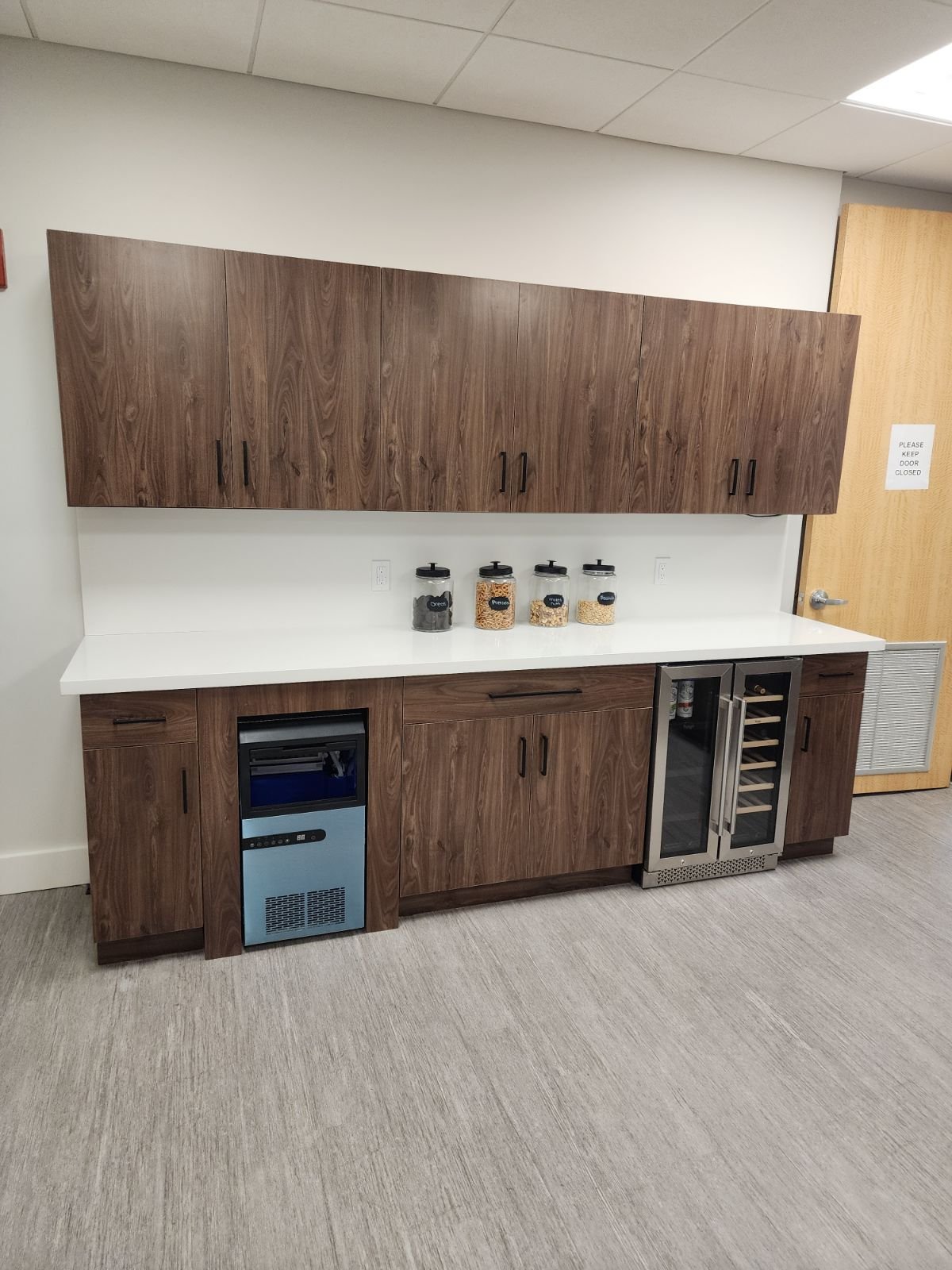 A kitchenette with wooden cabinets, a white countertop, and a small fridge with a glass door, along with four canisters on the counter and a sign on the door in the background.