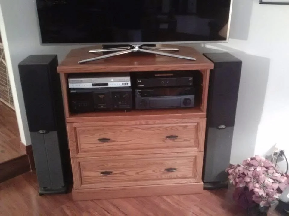 A wooden entertainment center holding a flat-screen TV, stereo components, and two large black speakers, with a pink flower arrangement on the floor nearby.