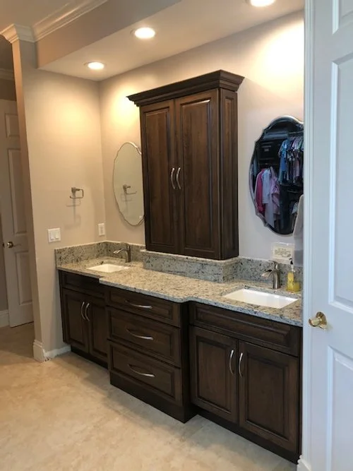 Bathroom vanity with granite countertop, two sinks, dark wood cabinets, and a mirror. Denim and purple clothing are visible in a hanging organizer on the wall.