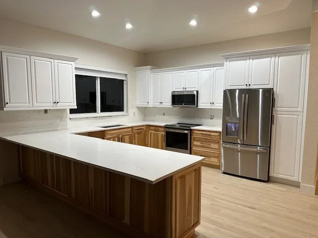 Kitchen with white upper cabinets, wooden lower cabinets, and stainless steel appliances including a refrigerator, microwave, and oven. An island with a white countertop is in the foreground.