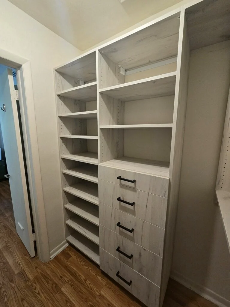 White built-in shelving unit with multiple open shelves and drawers, positioned against a wall in a room with hardwood flooring.