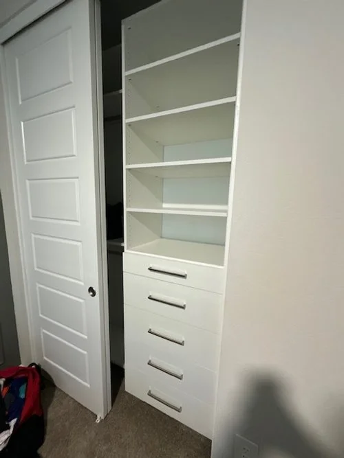 A white built-in bookshelf with five open shelves and four drawers at the bottom, next to a sliding door in a room with beige carpet.