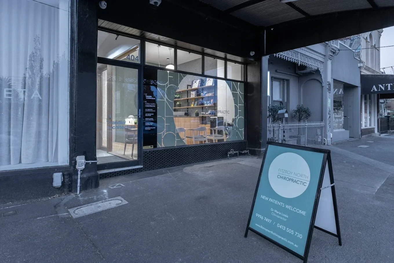 The storefront of a chiropractic clinic with a glass window and a sandwich board sign outside on the sidewalk promoting new patients welcome, with contact information and the name Fitzroy North Chiropractic.