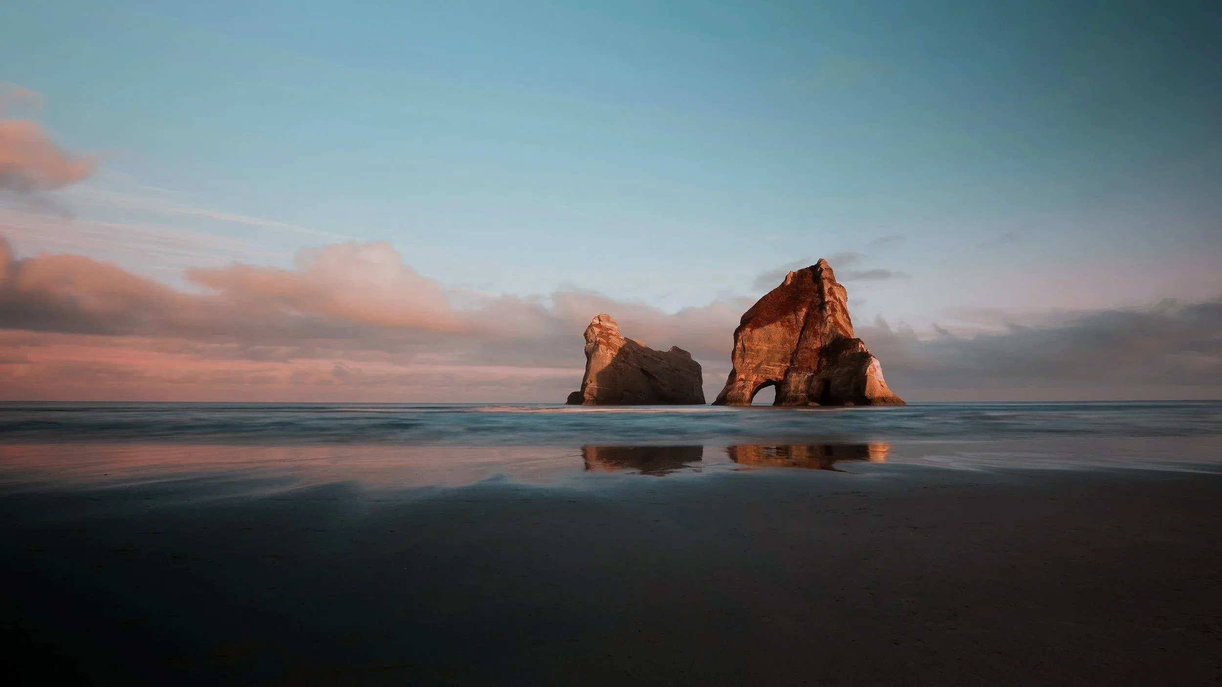 Two large sea stacks at the shoreline with gentle waves and a partly cloudy sky during sunset.