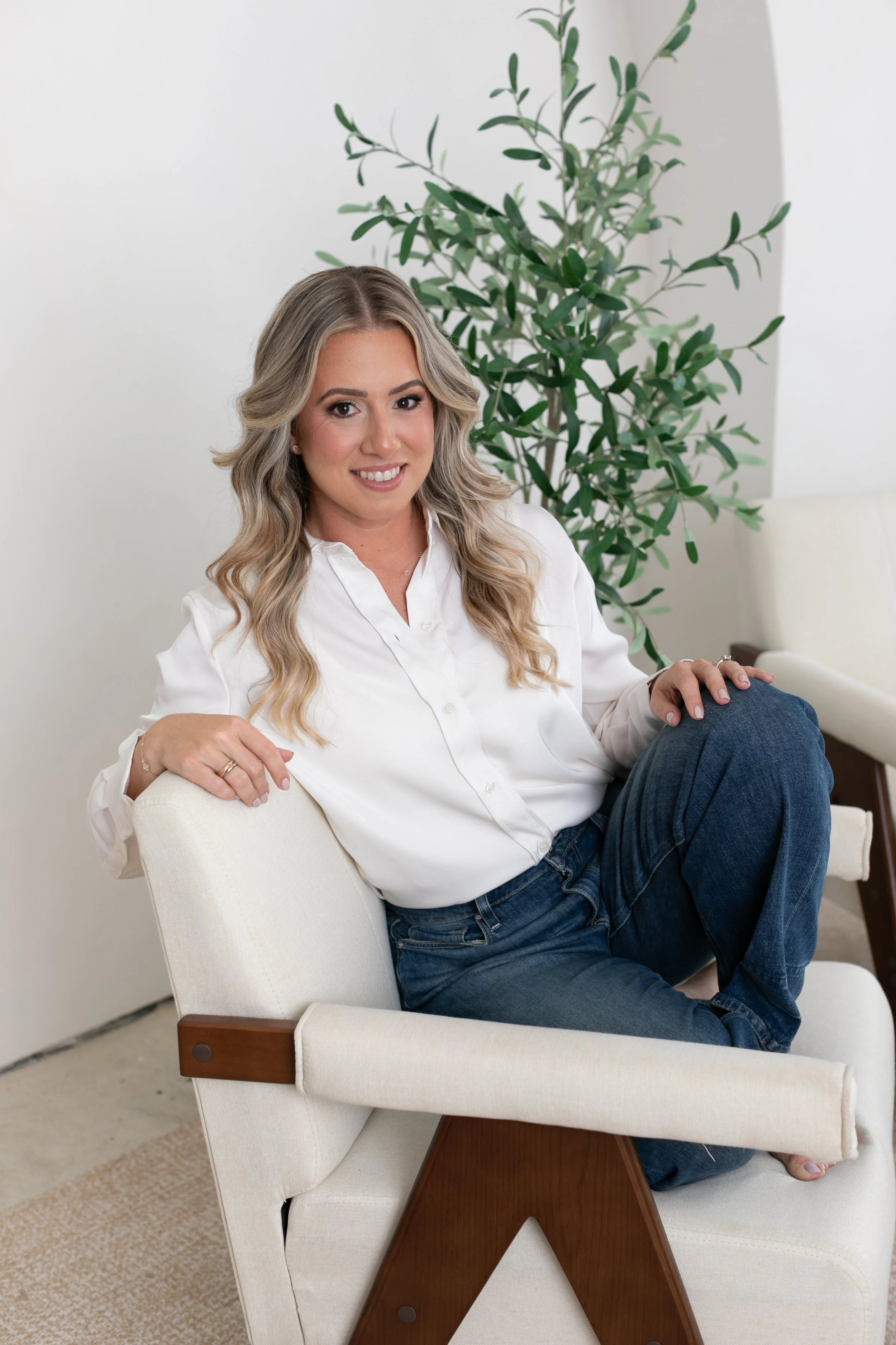 A woman with long wavy blonde hair, wearing a white button-up shirt and blue jeans, sitting on a modern cream armchair with wooden arms and smiling at the camera. There is a large green plant behind her.