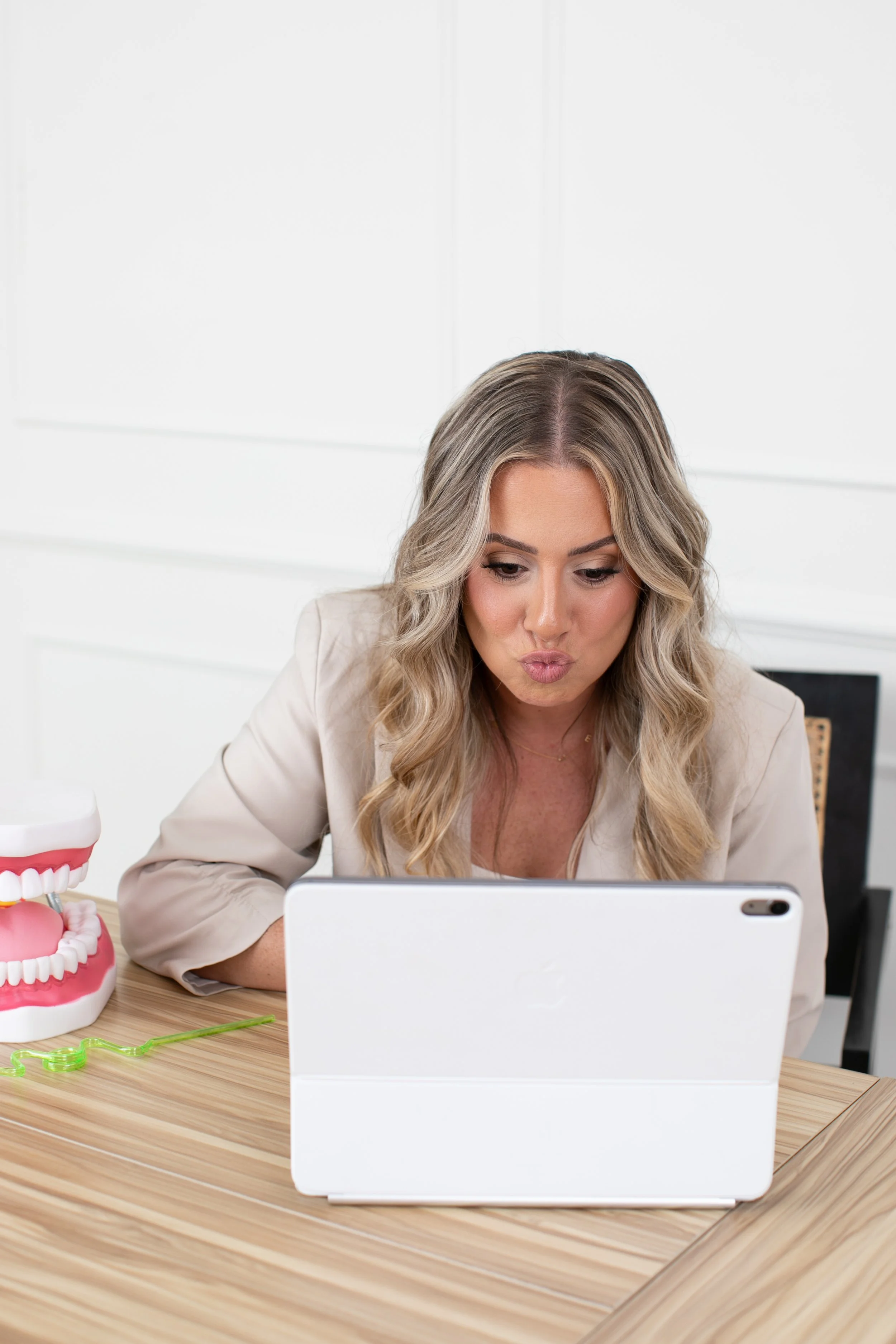 A woman with blonde wavy hair looking at a tablet on a wooden table, with a dental model of teeth and gums to her right.