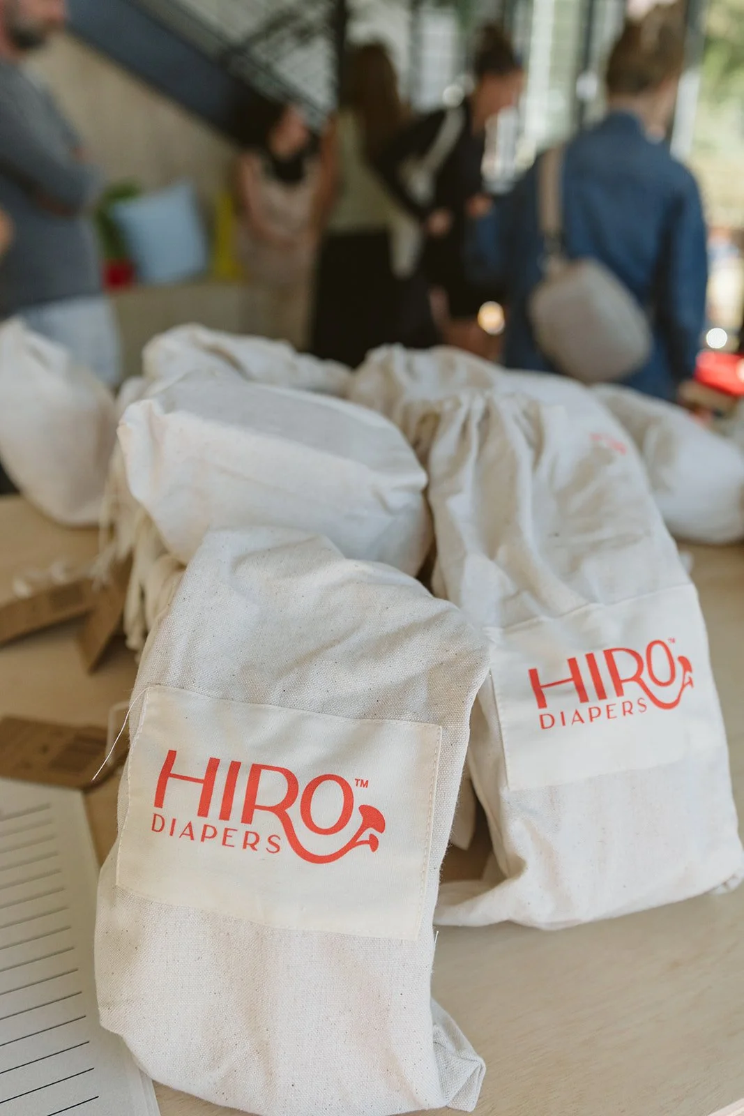 Cloth bags with red text reading 'HIRO DIAPERS' on a table, with people in the background at what appears to be a public indoor space.
