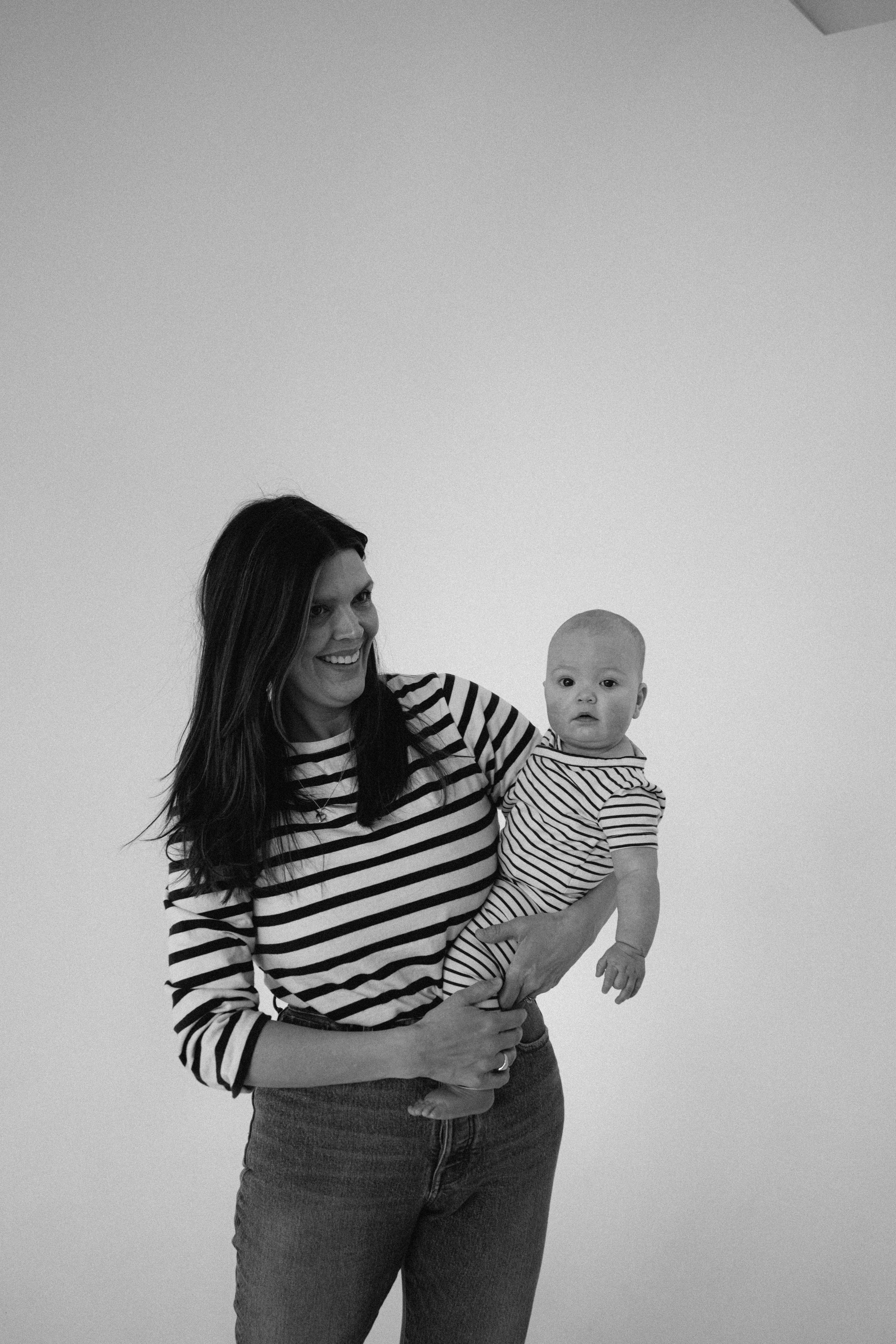 A woman smiling and holding a baby in her arms. Both are wearing striped shirts, with a plain background.