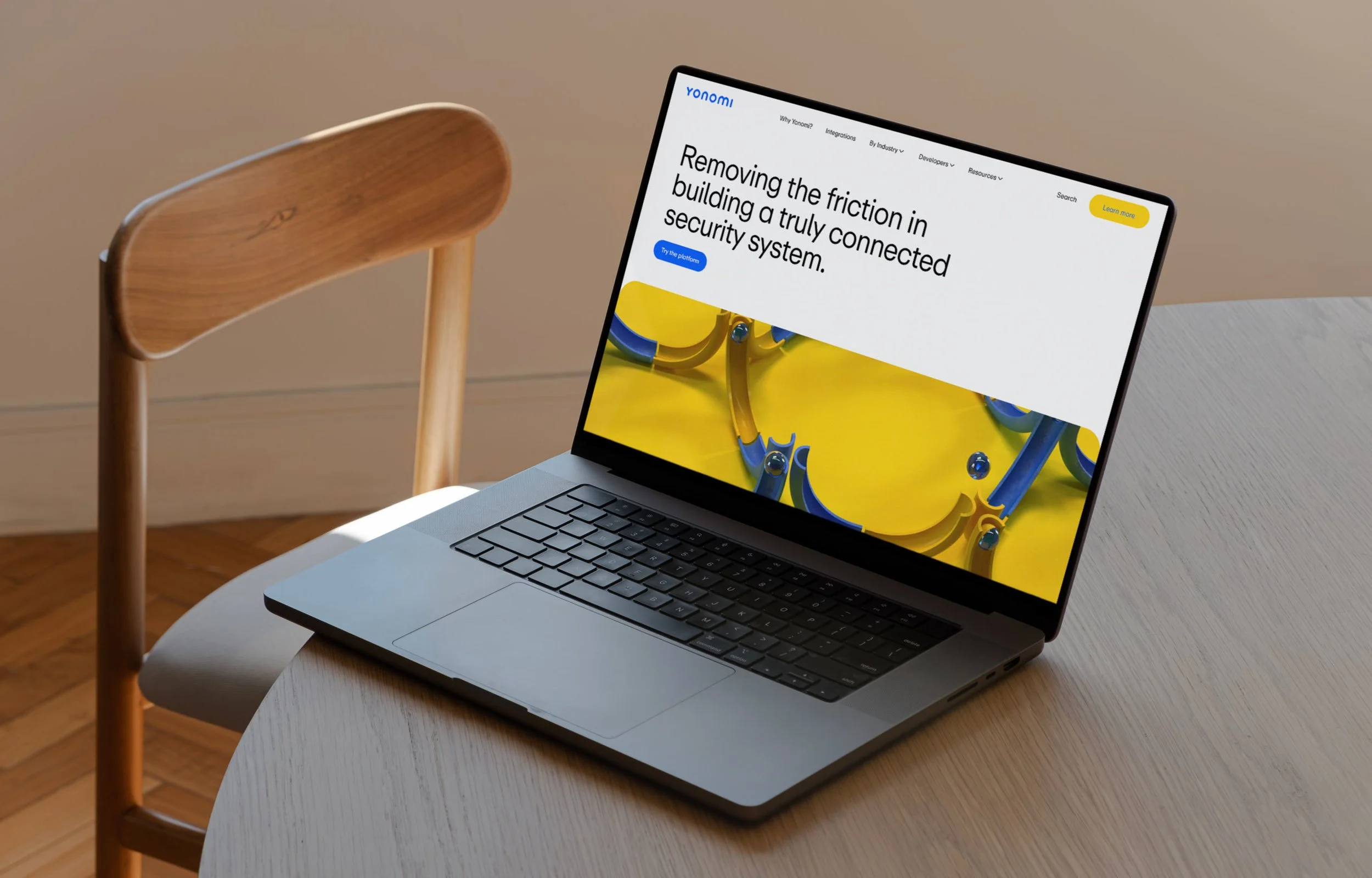 A laptop on a light wood table displaying a website with a white background and yellow, blue, and black accents, positioned next to a wooden chair in a room with light-colored walls.
