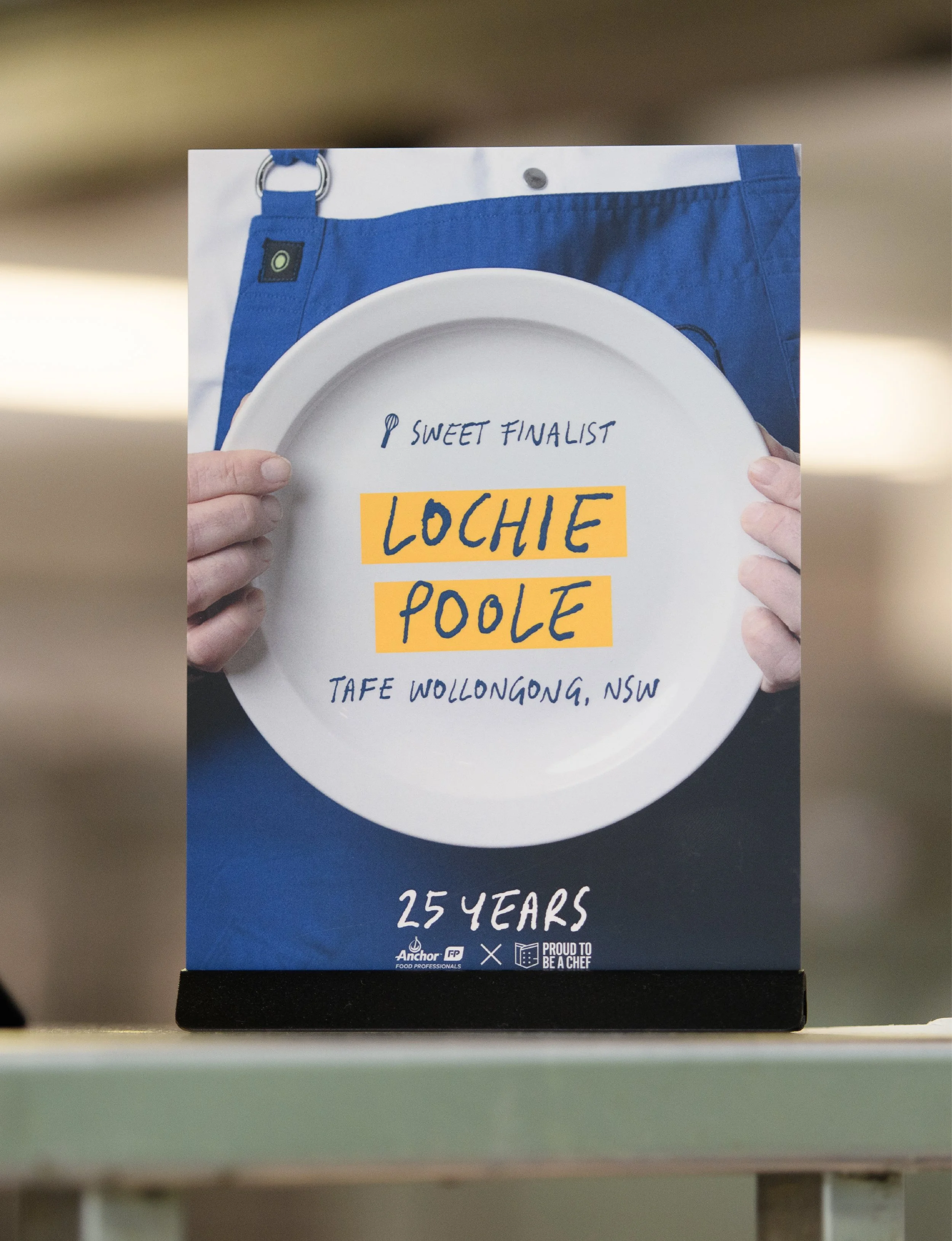 A person holding a white plate with handwritten text that reads 'Sweet Finalist Lochie Poole TAFE Wollongong, NSW'. The background shows a person wearing a blue apron.
