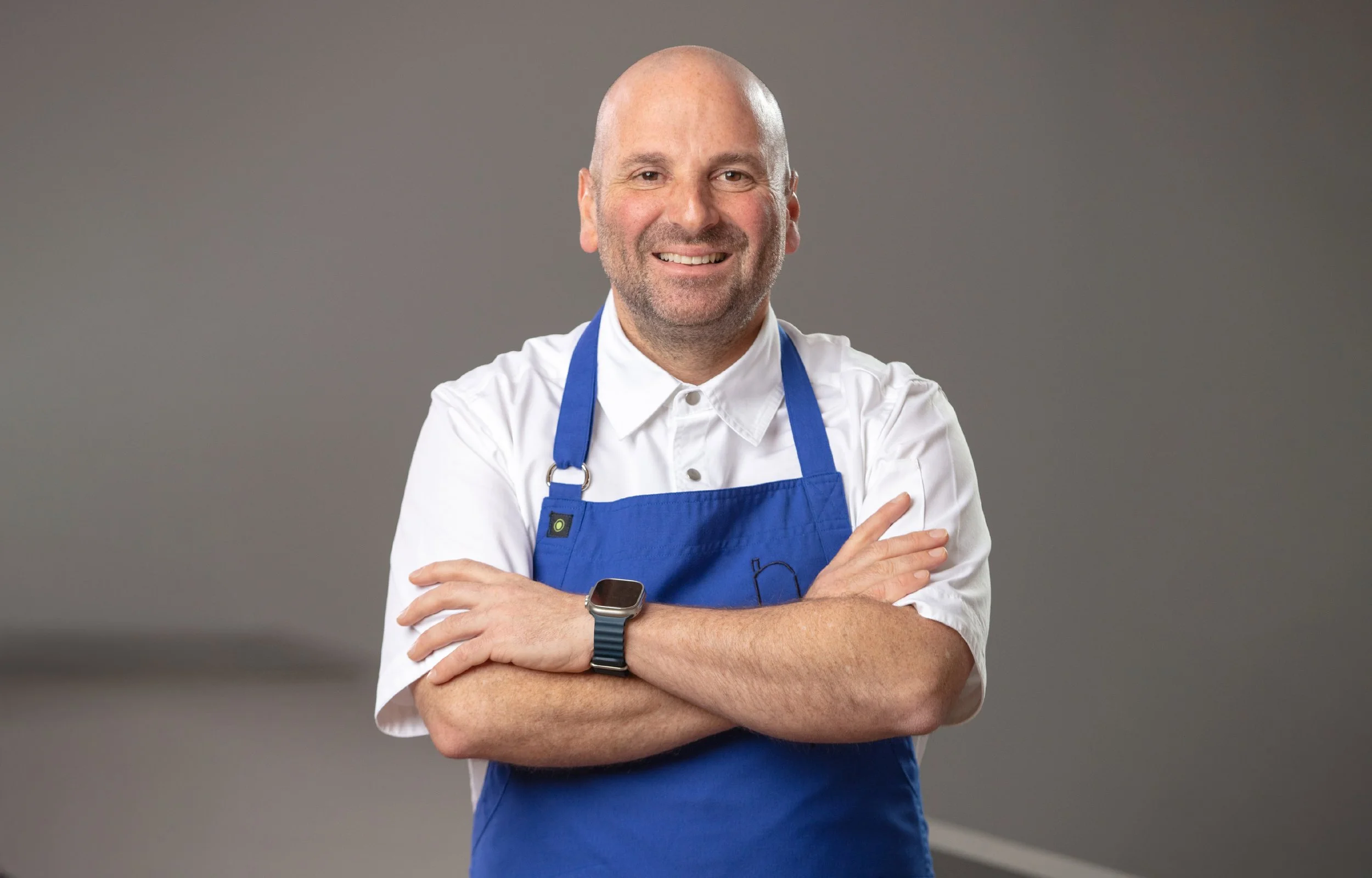 A chef smiling and wearing a white shirt, blue apron, and a smartwatch, standing with arms crossed against a gray background.