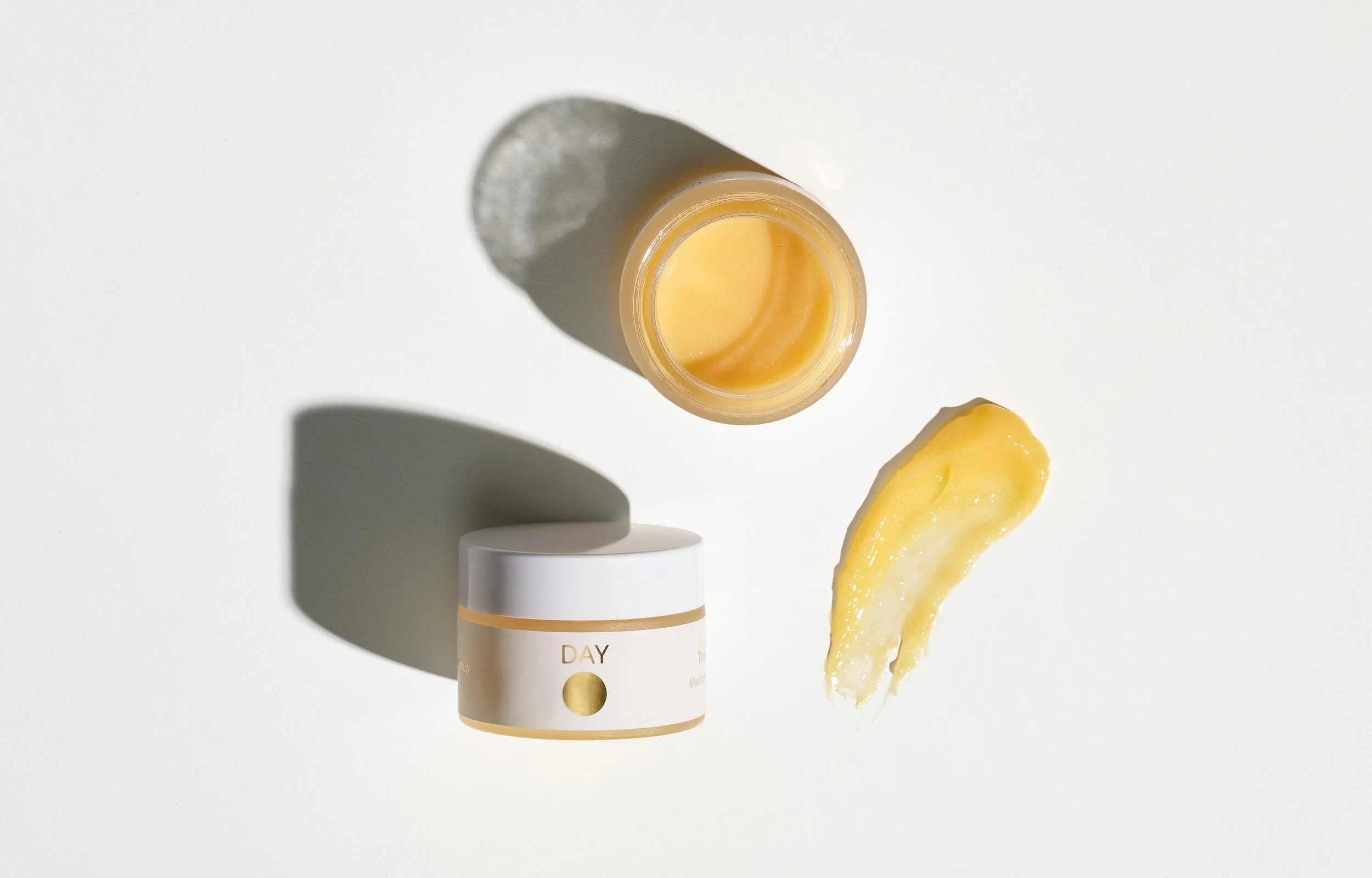A white jar labeled 'DAY' with a gold circle, an open small container with yellow cream inside and a swatch of yellow cream on a white surface, casting shadows.