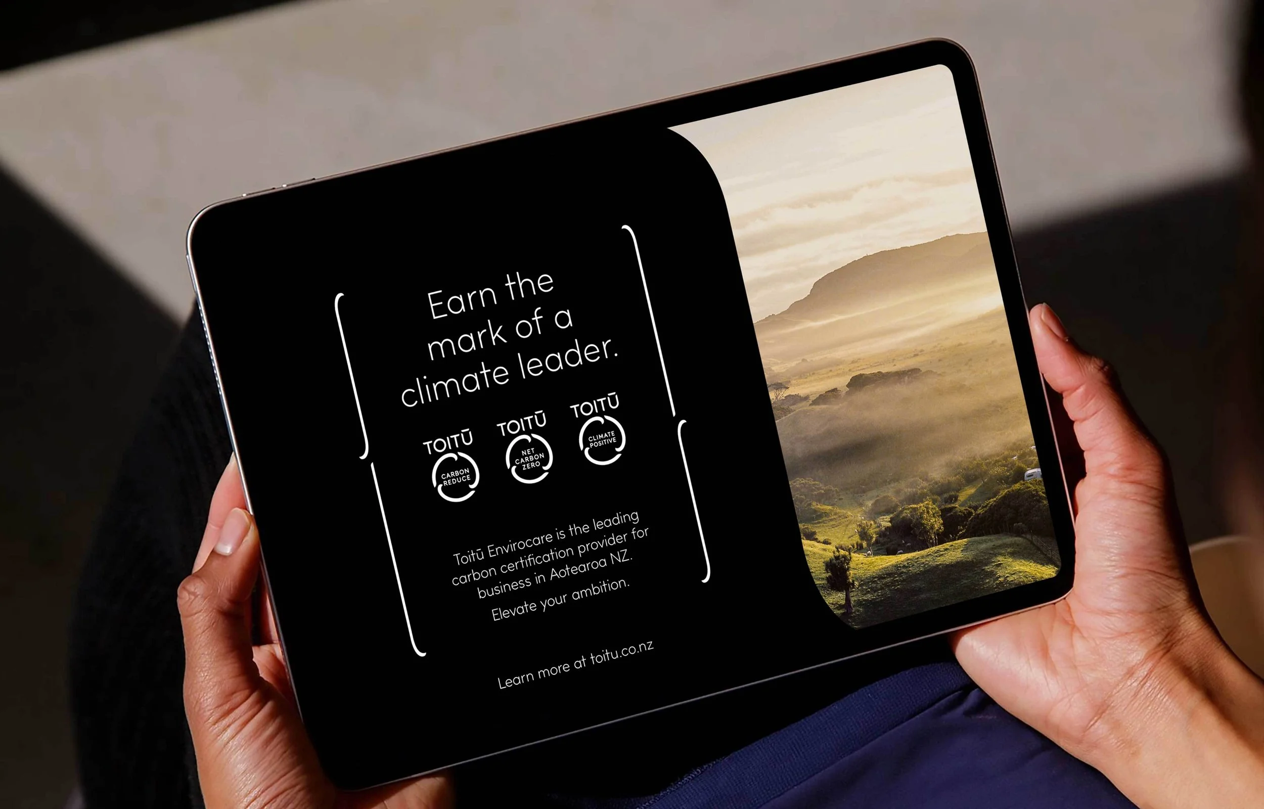 A person holding a foldable tablet displaying the Toitū environmental certification message with a scenic landscape in the background.