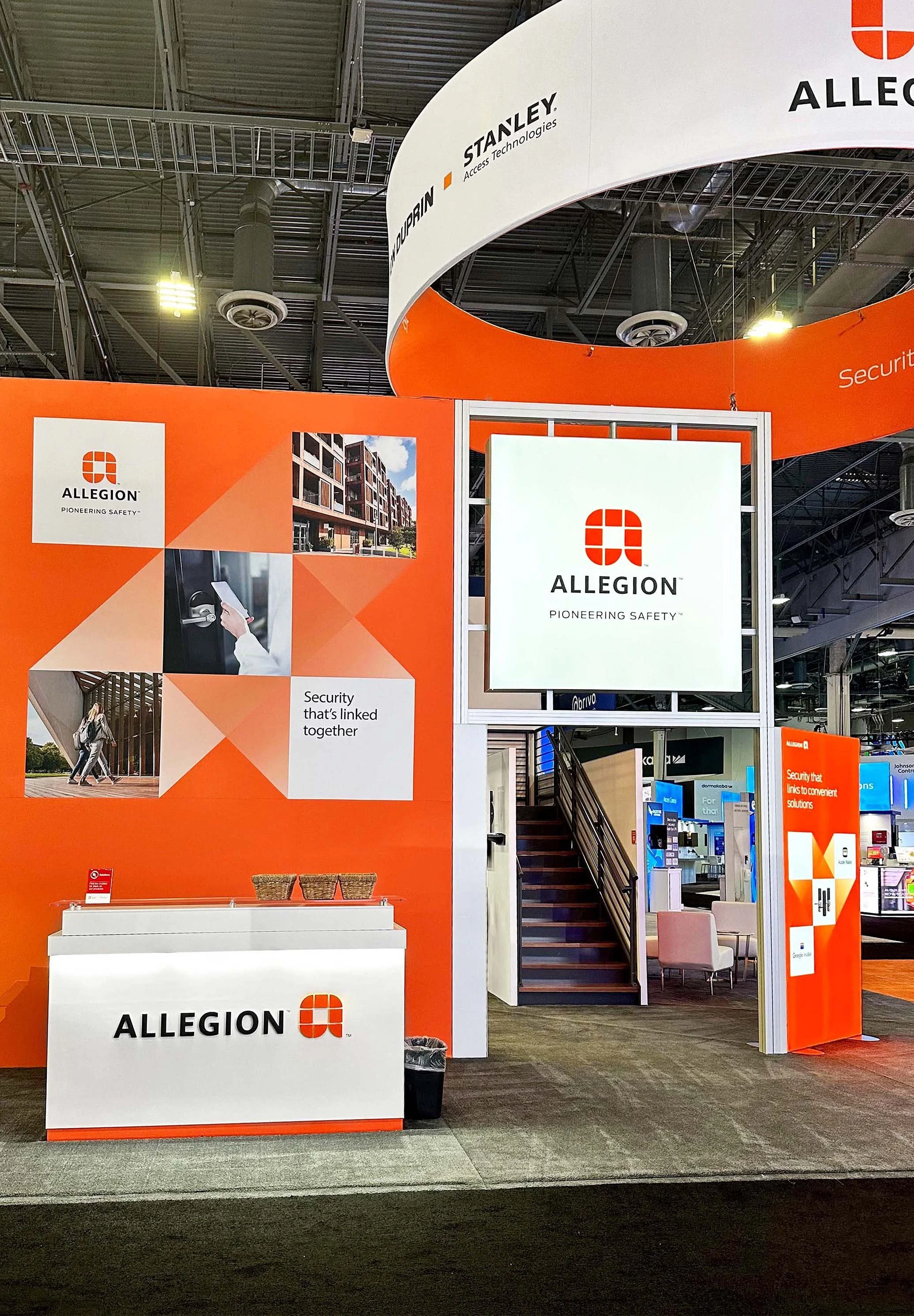 Allegion exhibit booth at a trade show with orange and white branding, displaying the Allegion logo, images of security products, and the slogan 'Pioneering Safety.'