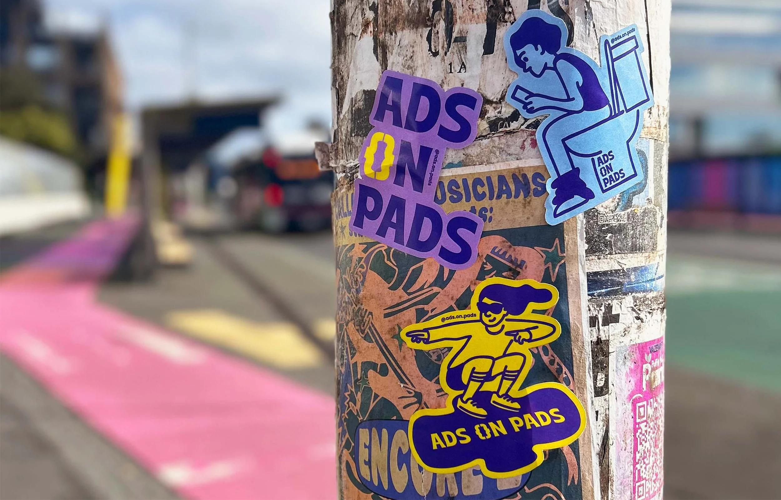 Close-up of a weathered pole covered in colorful stickers, including ones that say 'ADS ON PADS' and feature a cartoon person sitting on a toilet and a jumping person, with a busy street and pink and yellow painted crosswalk in the background.