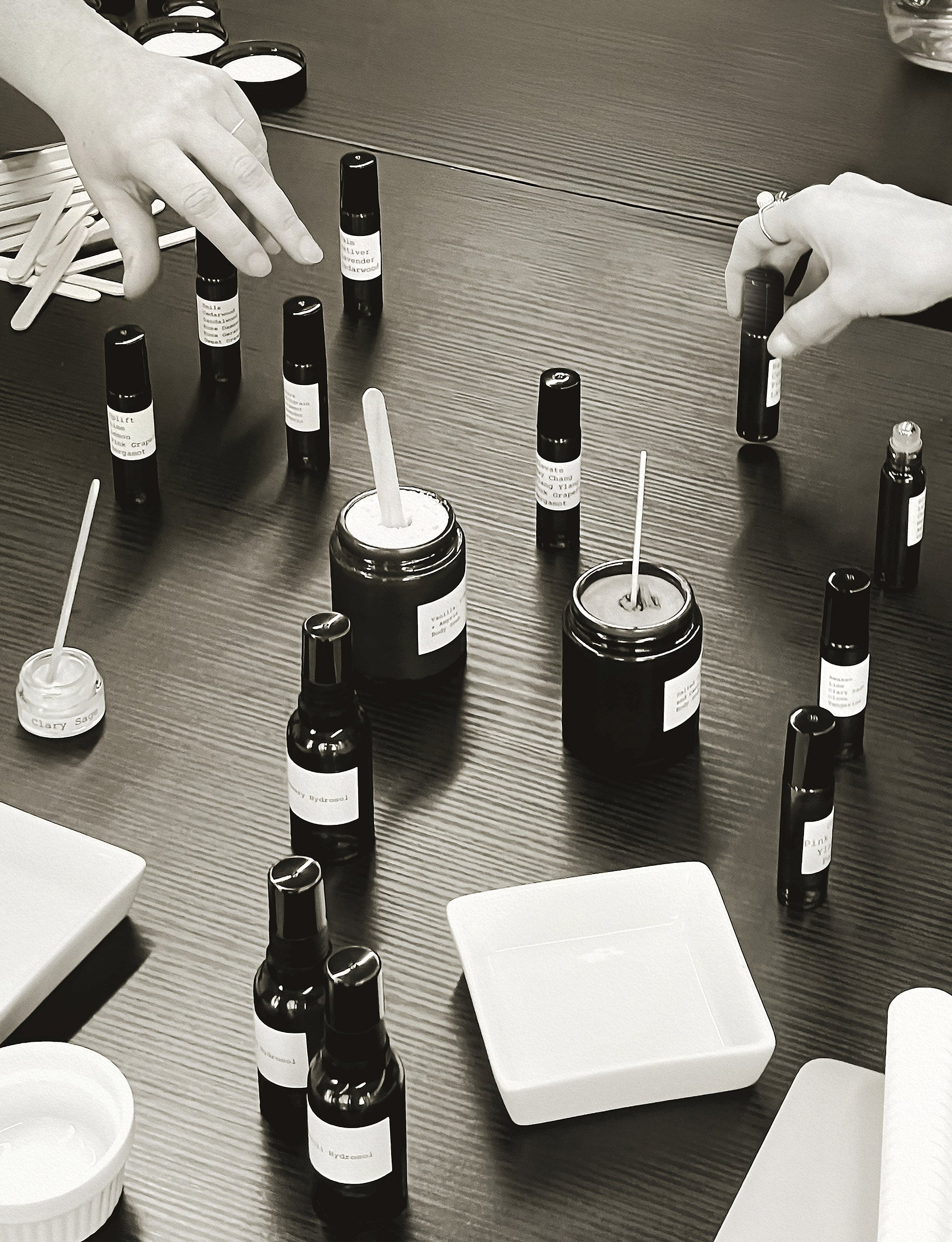 A table with various bottles of essential oils, some with droppers and small containers, with two hands reaching for bottles.