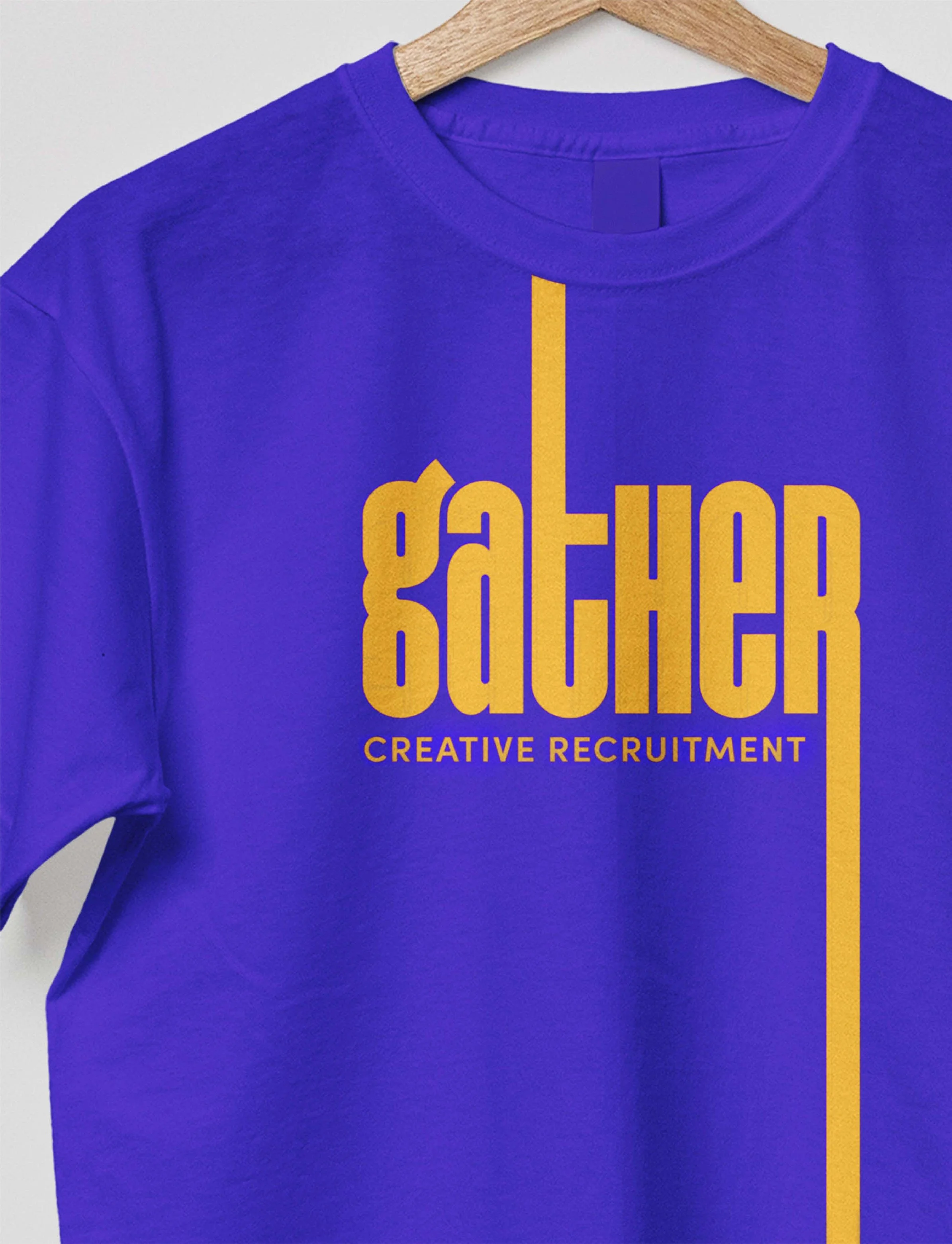Purple T-shirt hanging on a wooden hanger with yellow text that says 'GATHER' and smaller text below that says 'CREATIVE RECRUITMENT'.