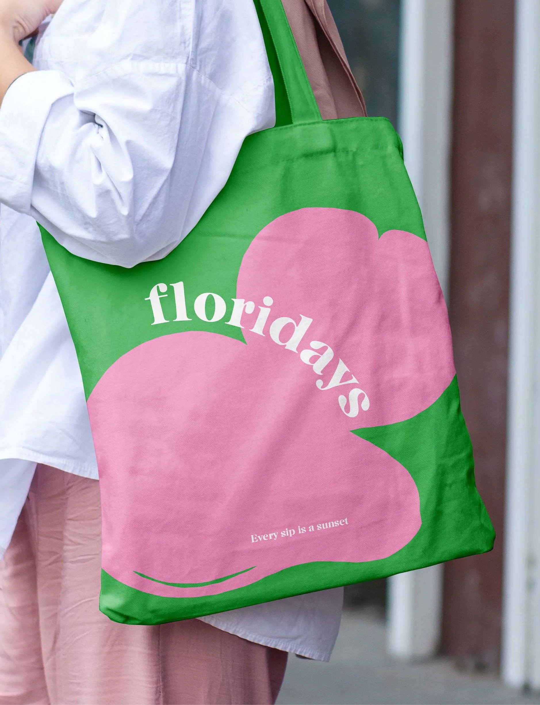 A person wearing a white shirt and pink pants carrying a green tote bag with pink peach shapes and the word 'floriday' written on it, along with the phrase 'Every sip is a sunset'.
