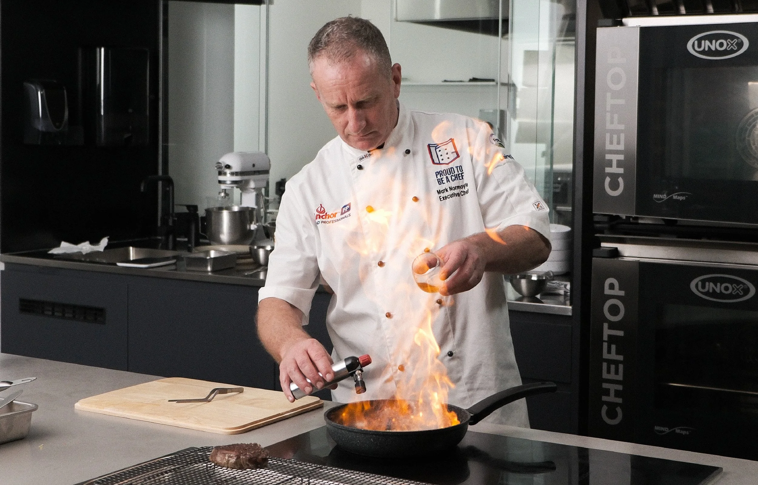 Chef in a white uniform stands in a commercial kitchen, igniting a flambé in a skillet with a blue flame and a burst of fire.