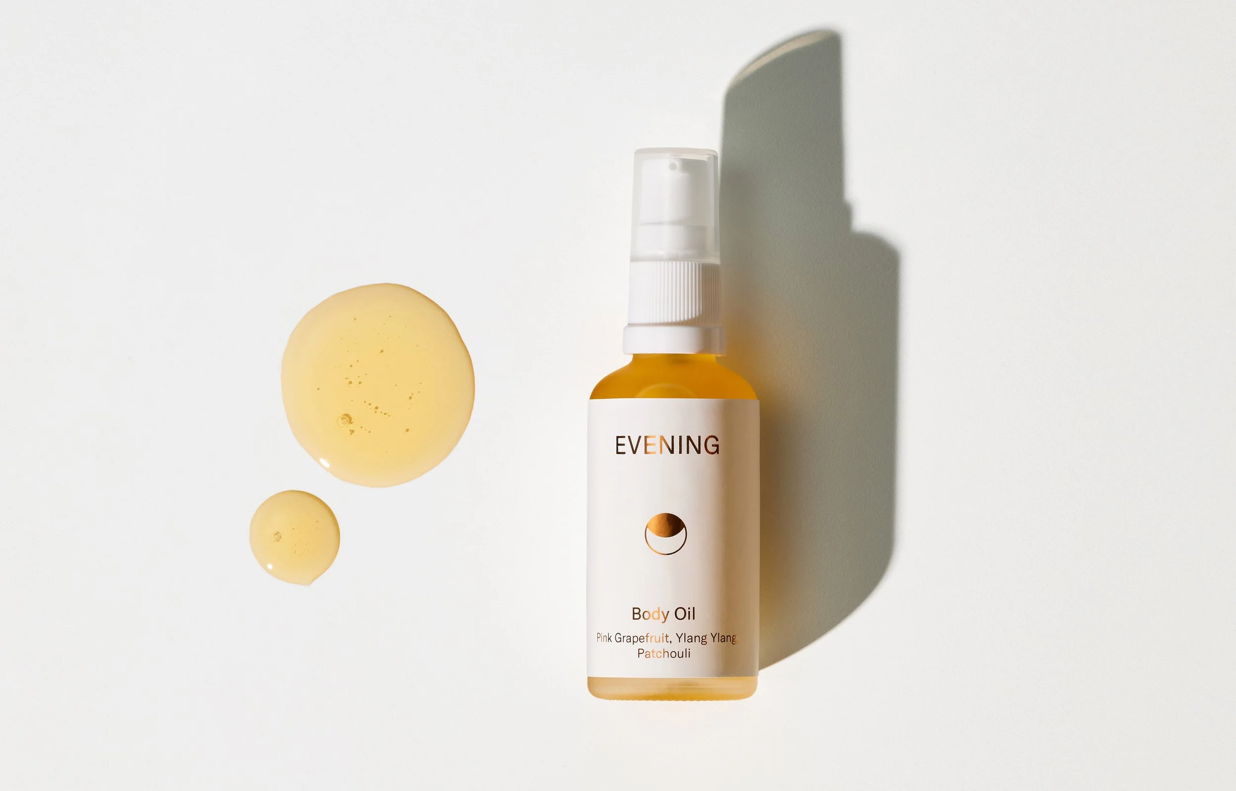 Bottle of evening body oil with droplets of oil on a white surface, shadow cast behind the bottle.