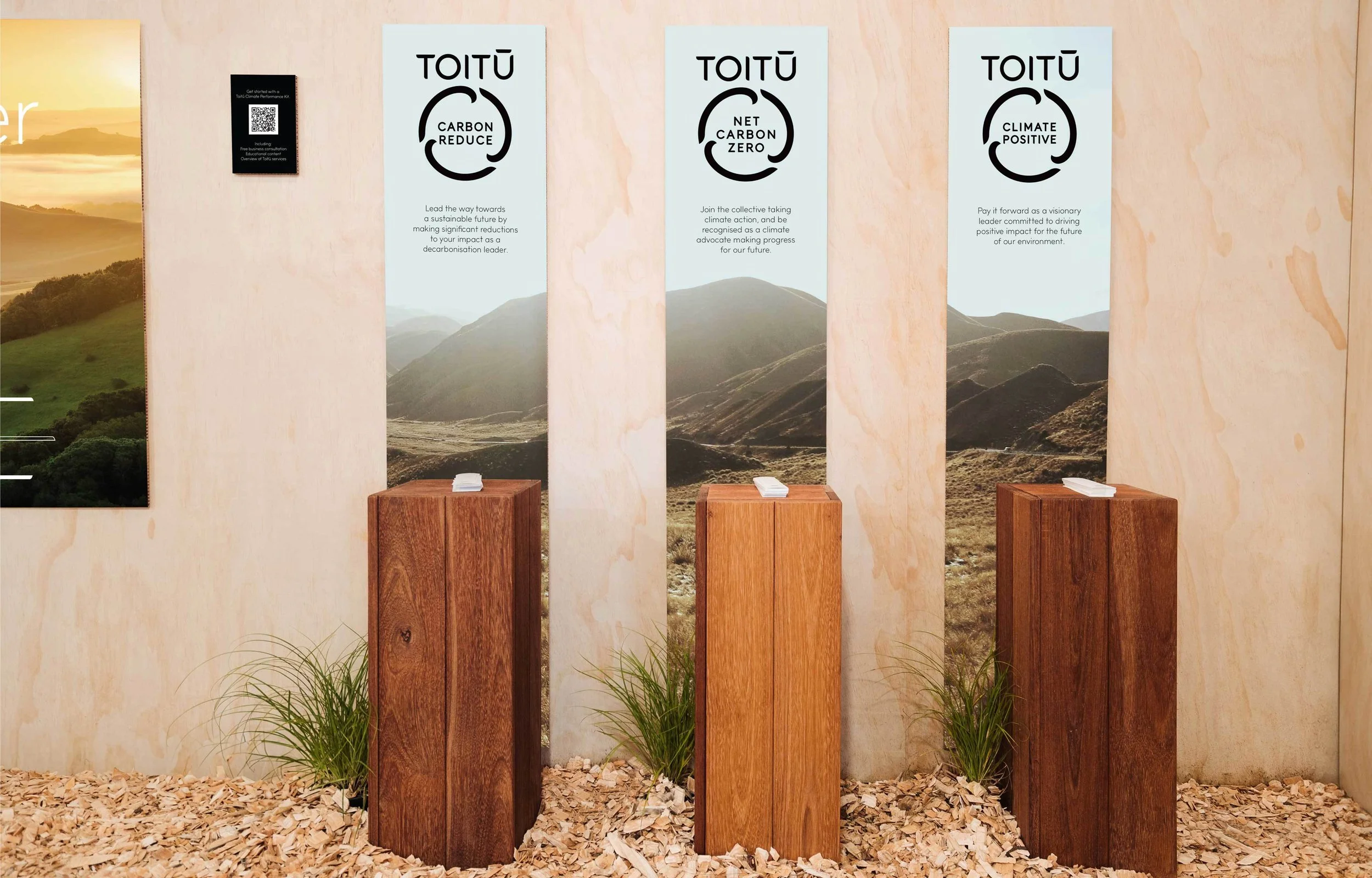 Three wooden pedestals in front of a scenic nature background with mountains and grassy landscape. Behind them are three vertical banners with Toitū messages about reducing carbon emissions, net zero carbon, and climate positivity.