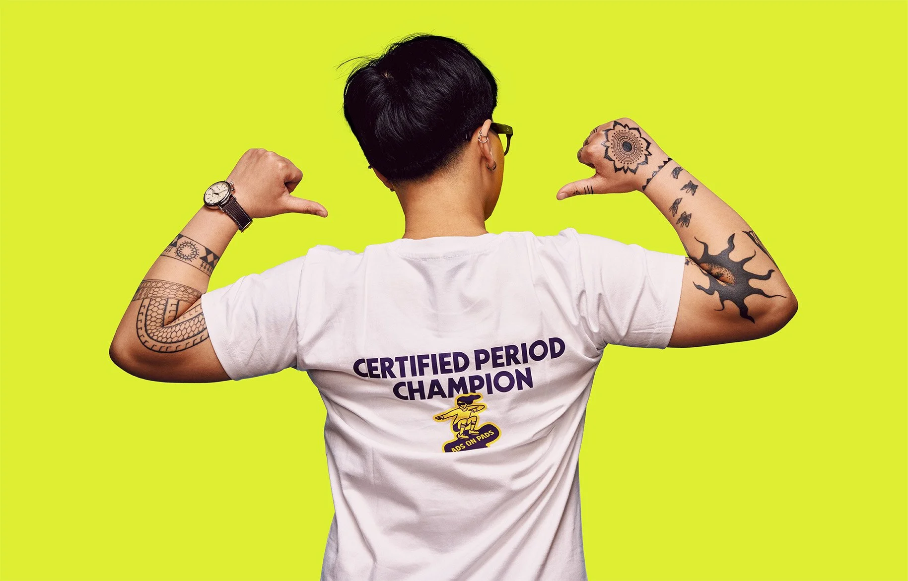 A woman with short dark hair, glasses, and tattoos on arms, flexing muscles from behind, wearing a white t-shirt with text 'Certified Period Champion' and a graphic of a person on pads, against bright yellow background.
