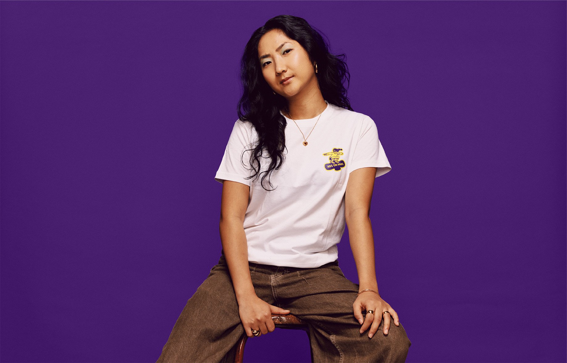 A woman with long dark hair, wearing a white T-shirt with a logo, brown pants, and jewelry, seated on a stool against a purple background.