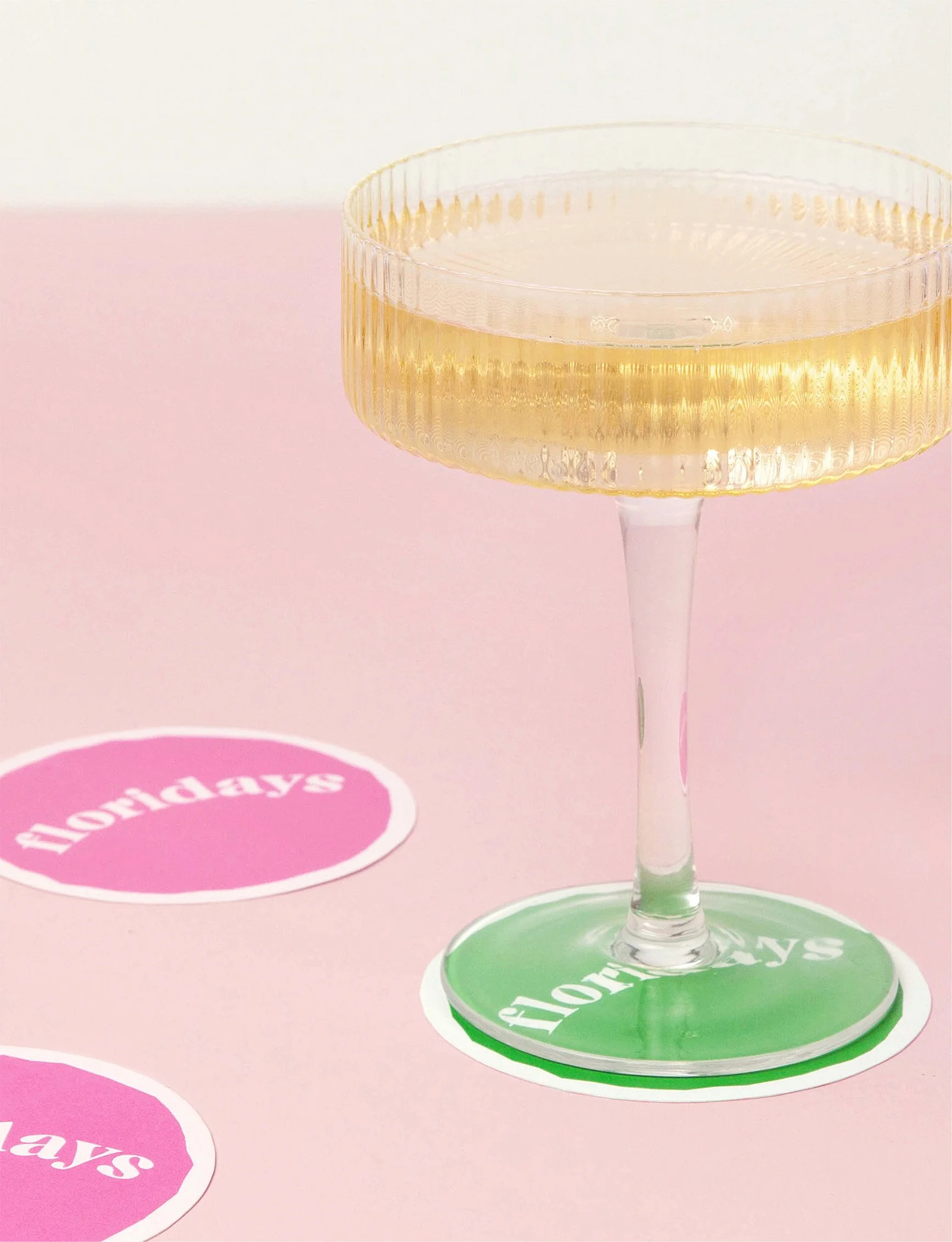 A vintage champagne coupe glass with a gold tint sitting on a pink surface with pink and green coasters labeled 'holidays'.