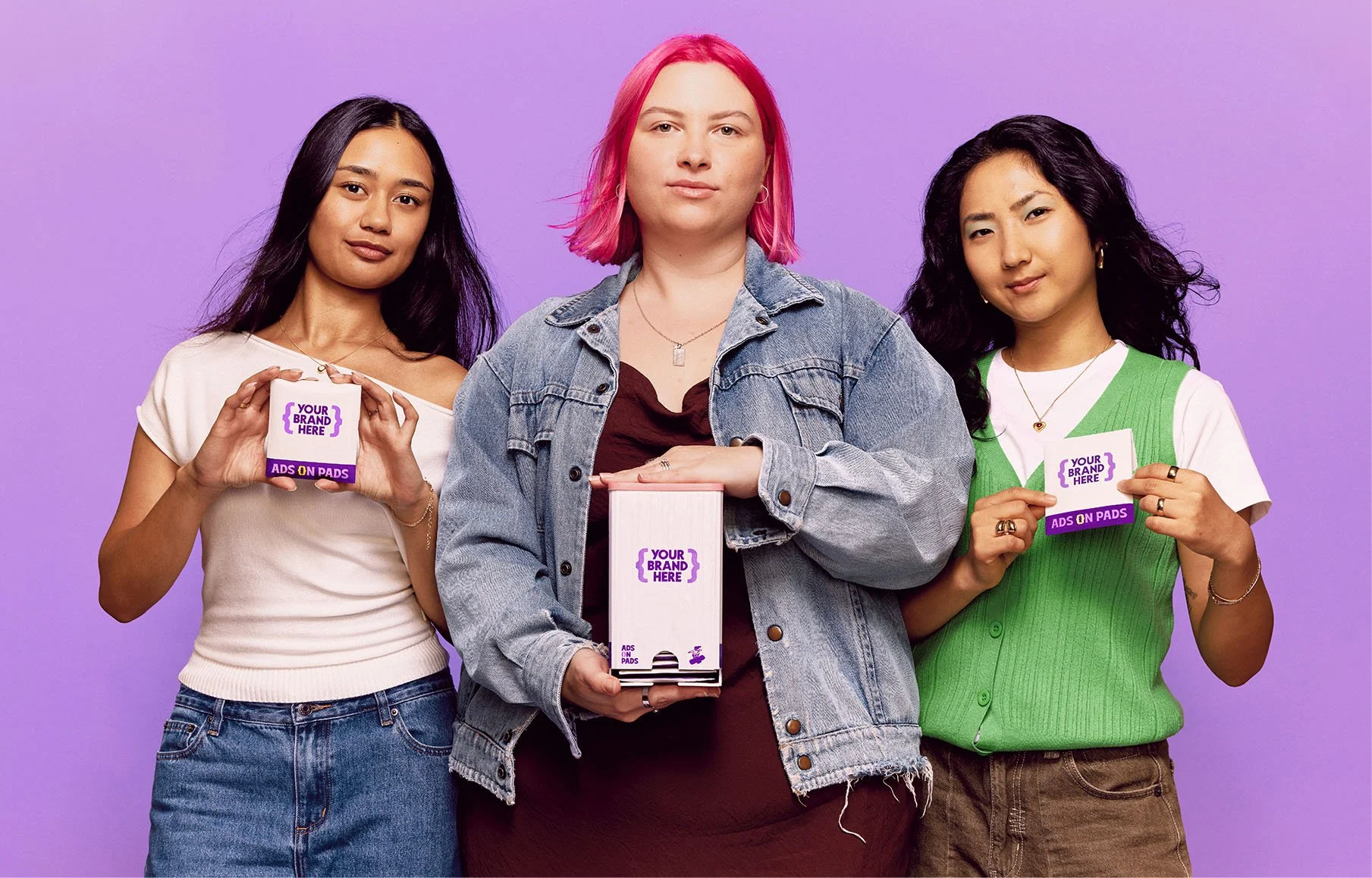 Three women posing against a purple background, holding promotional display cards and a box for ads on pads, with one in the center wearing a denim jacket and pink hair.