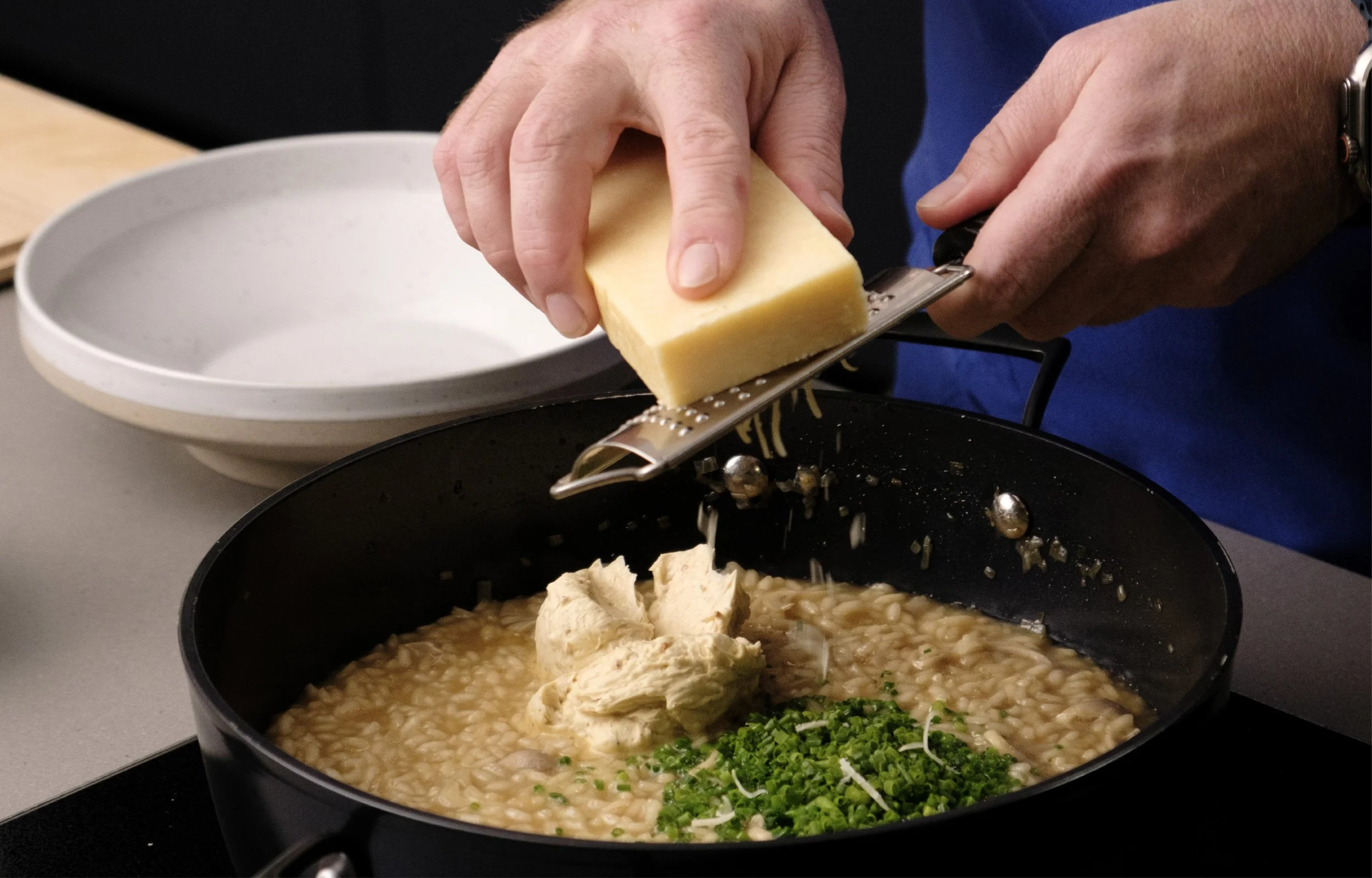 A person grates cheese into a pot of cooked risotto with green herbs. The person is holding a block of cheese and a cheese grater. There is a white plate in the background.