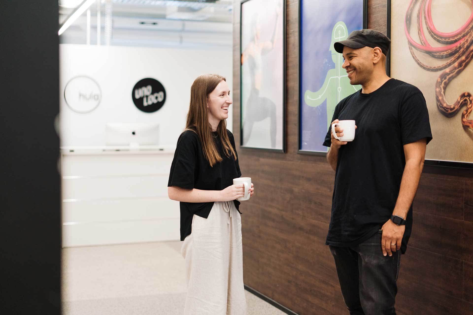 A man and woman smiling and talking while holding coffee mugs in an indoor office or lobby area, with artwork on the wall behind them.