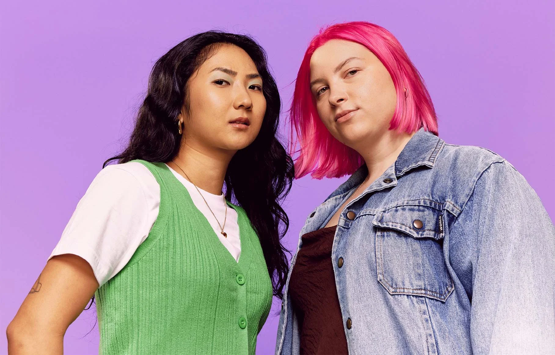 Two women standing close together against a purple background, one with long dark hair in a green vest and white shirt, the other with bright pink hair in a denim jacket.