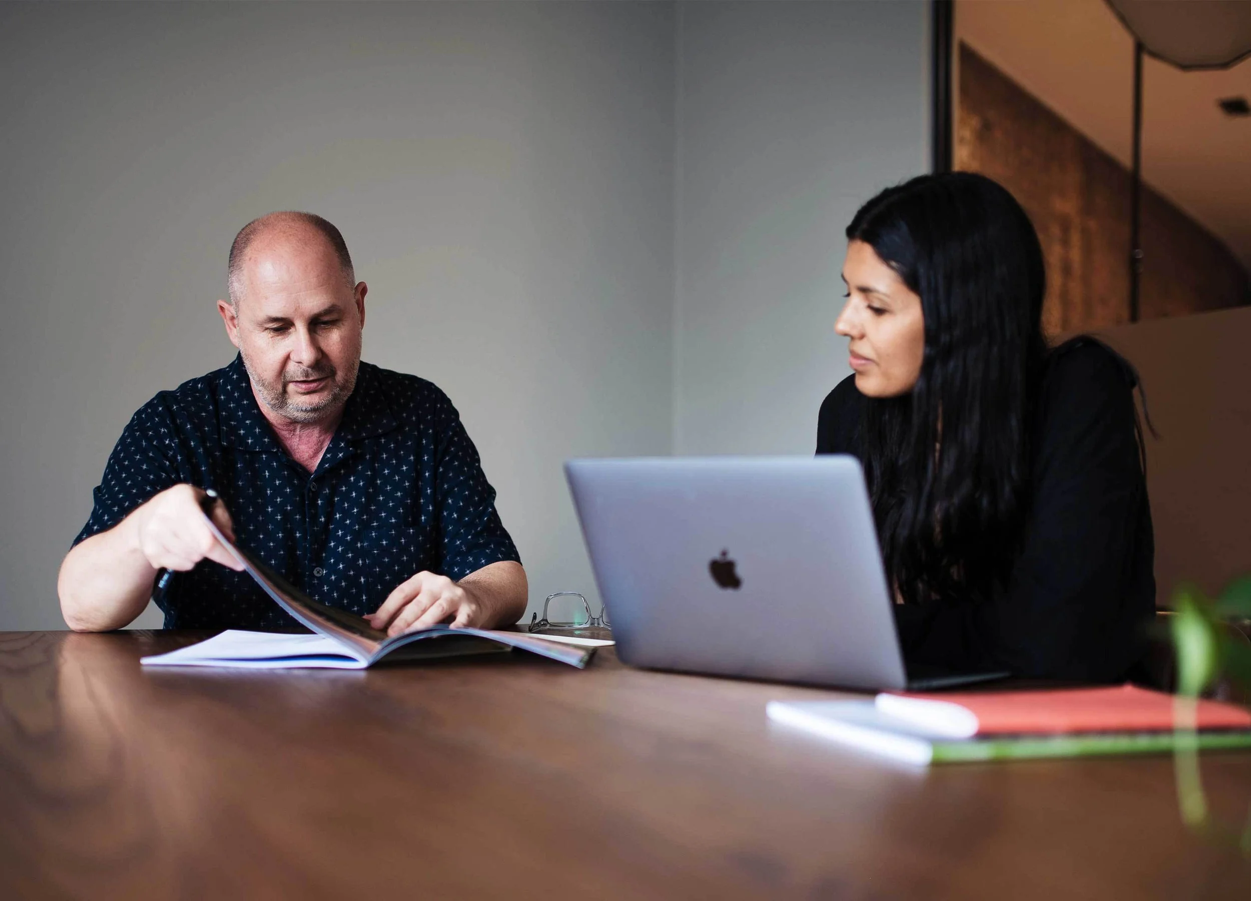 Two people sitting at a wooden table reviewing documents and working on a laptop in a modern office.