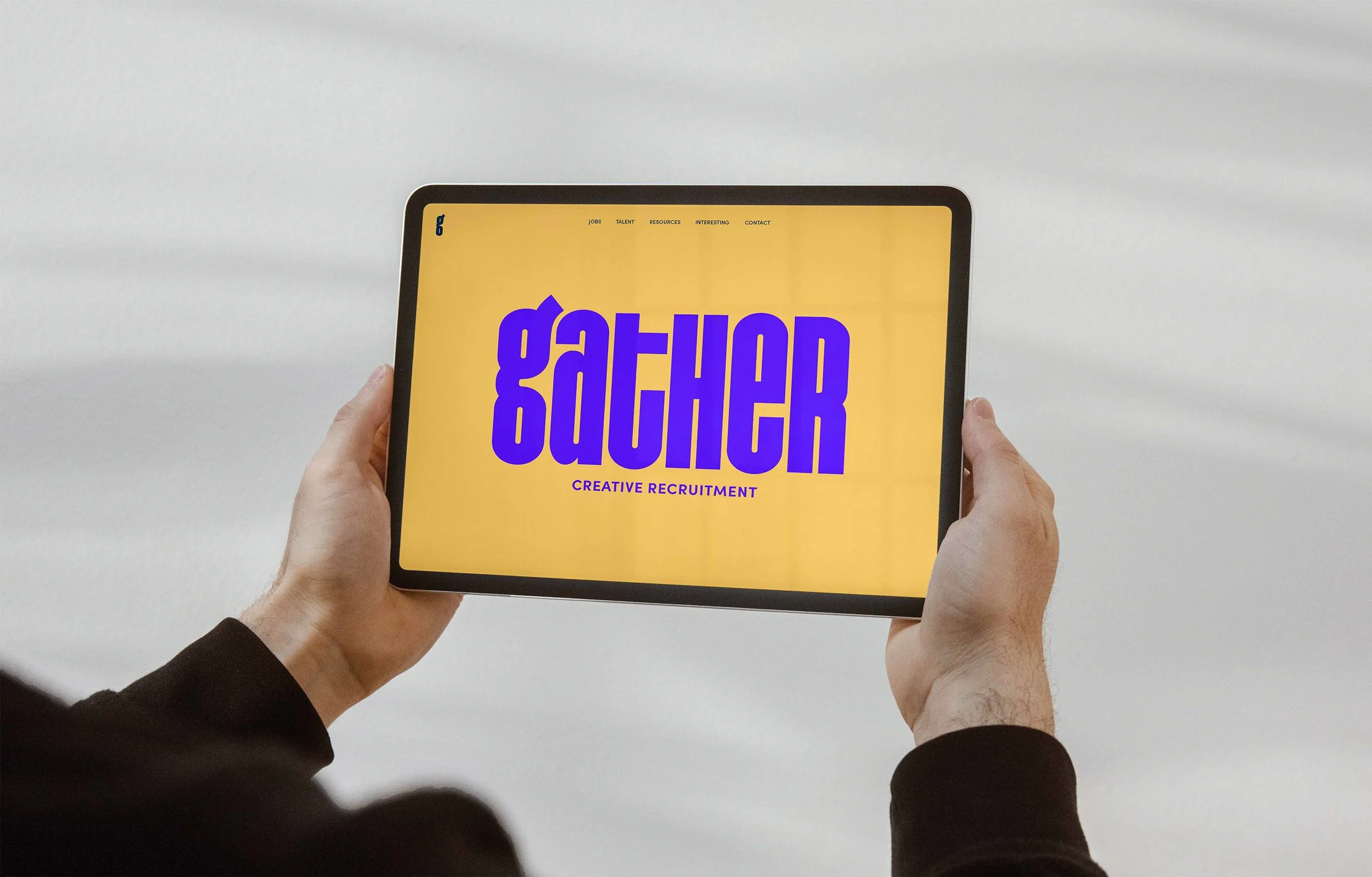 Person holding a tablet displaying a website with the word 'GATHER' in large purple letters, and the phrase 'CREATIVE RECRUITMENT' underneath, on a yellow background.
