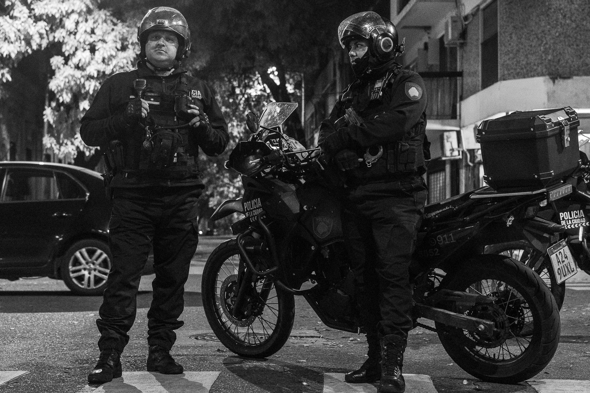 Dos policías patrullando en la calle durante la noche, uno con motocicleta y ambos con cascos y uniformes negros.