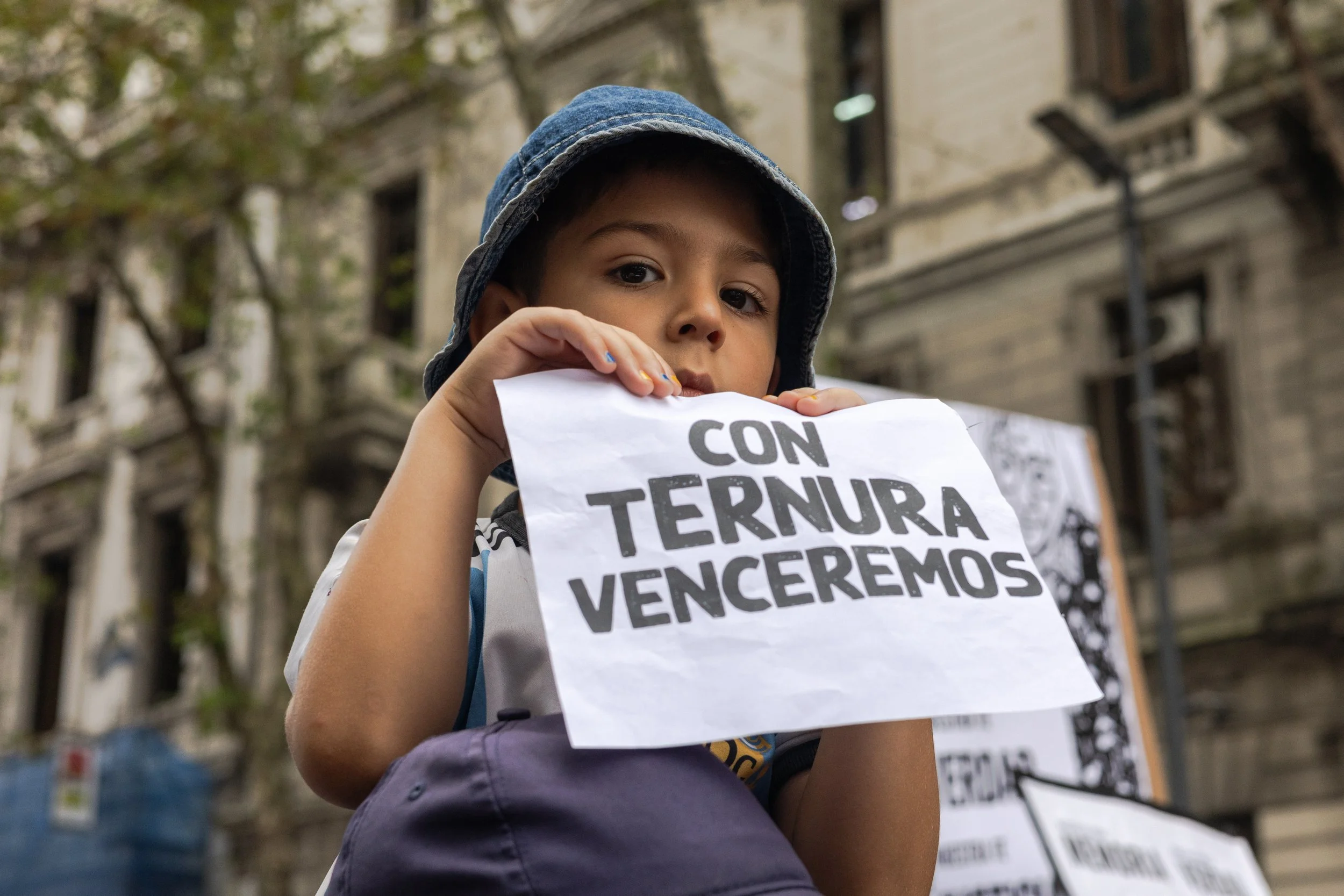 Un niño sostiene un cartel que dice 'Con ternura venceremos' durante una protesta en la calle con edificios antiguos de fondo.